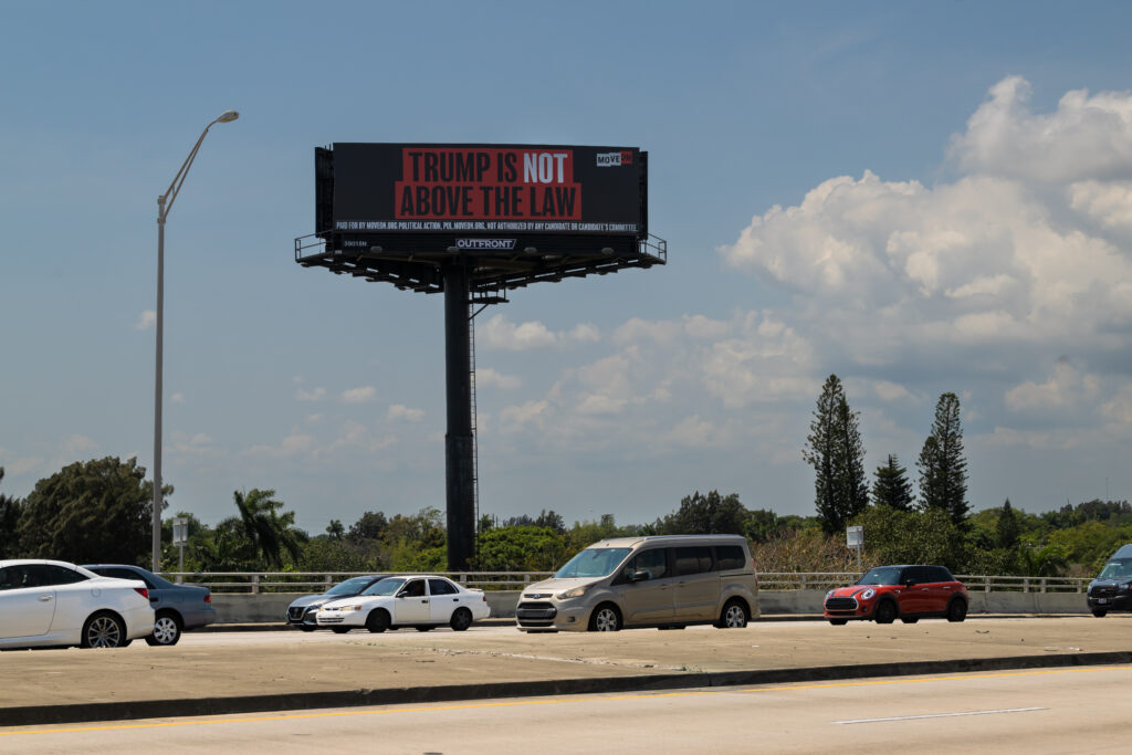 A large MoveOn billboard reads: "Trump is not above the law" against a blue sky with clouds and over the highway approaching Mar-a-Lago, Florida where many cars are seen lined up. 