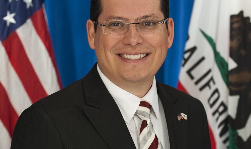 Headshot of Rudy Salas smiling in a black blazer and a striped red, white, and grey tie.