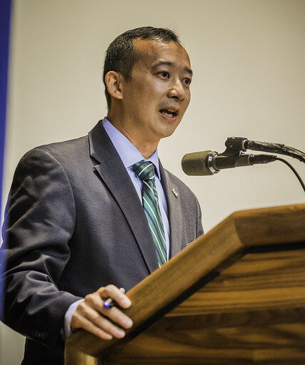 Lanon Baccam giving a speech in a grey suit, blue shirt and green striped tie while casually holding a pen in his right hand.