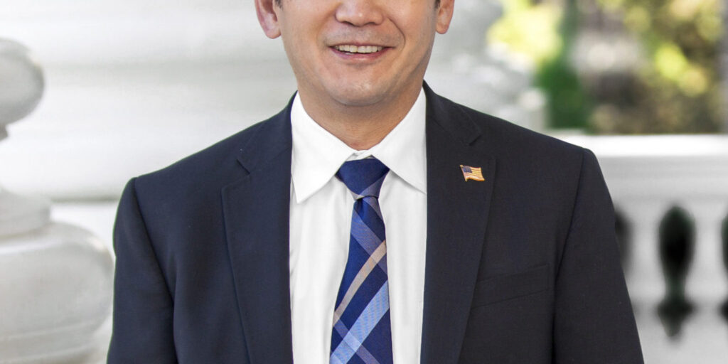 Headshot of Dave Min in a dark suit with a striped blue tie and white shirt.