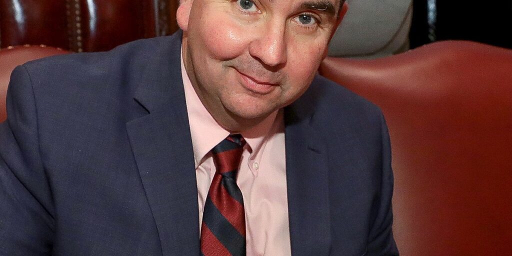 John Mannion in a blue blazer smiling at the camera while signing a document.
