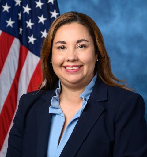 Headshot of Dr. Yadira Caraveo in a dark blue blazer and a light blue blouse with an American flag and blue background.