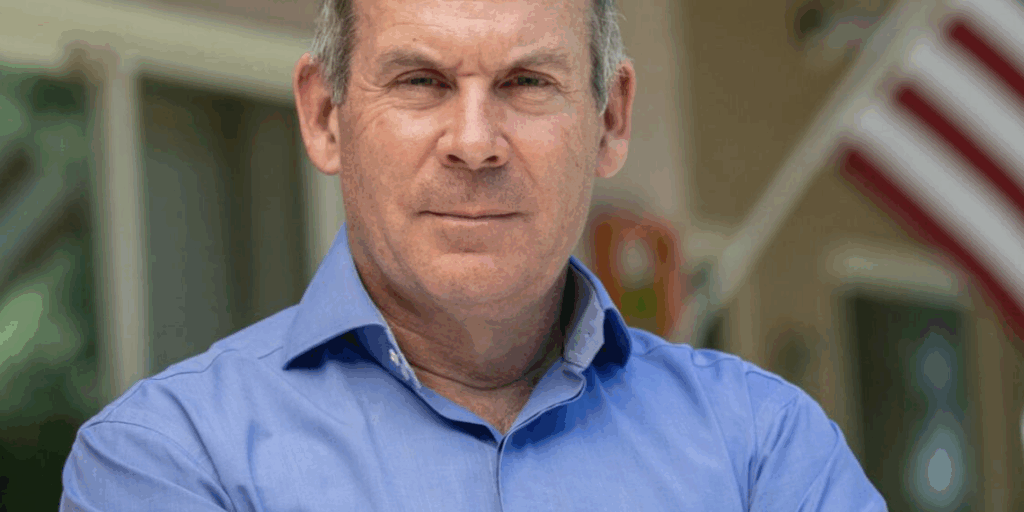 Adam Frisch wearing a light blue collared button up shirt crossing his arms and gazing into the camera.