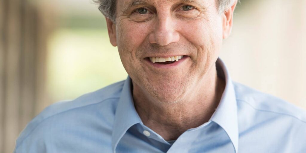 The image shows Sherrod Brown smiling warmly while wearing a light blue button-down shirt. His hair is slightly tousled, and the background is softly blurred, giving the image a relaxed and approachable feel.