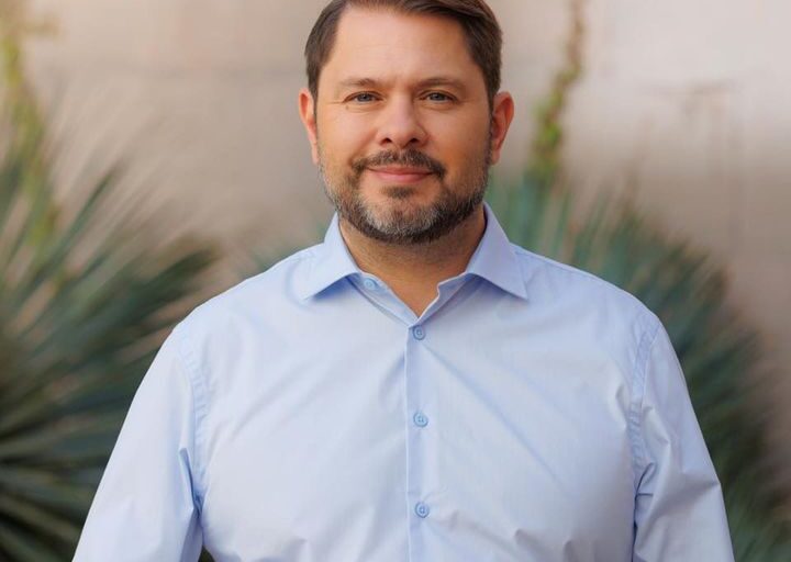 Ruben Gallego stands confidently in a light blue shirt with a desert landscape in the background.