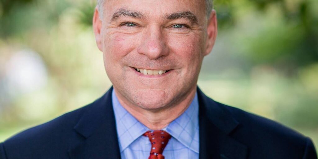 Tim Kaine in blazer with a red tie on in front of a blurred greenery background.