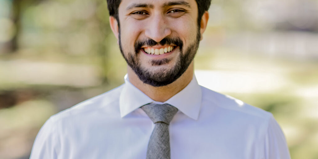 Greg Casar is shown smiling outdoors, wearing a white dress shirt and gray tie, with a blurred natural background.