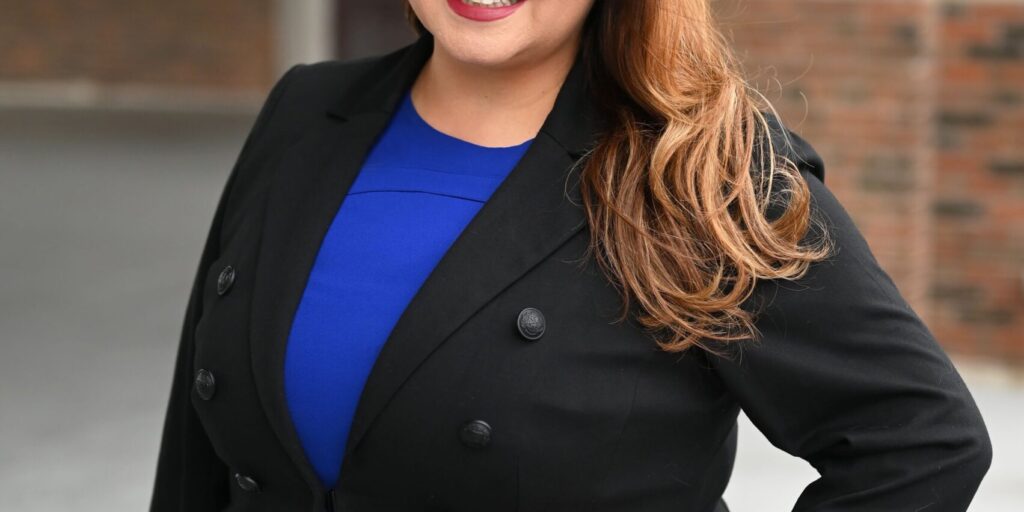 Delia Ramirez is pictured smiling confidently in this image, wearing a blue dress and a black blazer, standing with her hand on her hip in front of a building with arched windows.