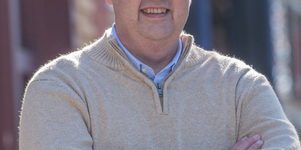 Curtis Hertel smiling confidently with arms crossed, wearing a beige sweater, and standing in front of a blurred street scene with buildings and autumn trees in the background.