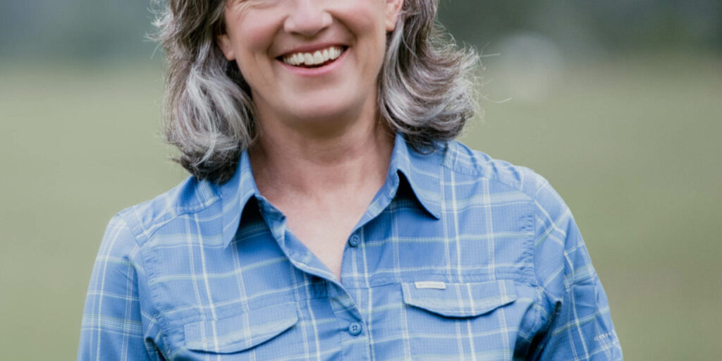 Monica Tranel is smiling outdoors in a blue plaid shirt against a blurred natural background, embodying her down-to-earth and approachable nature.