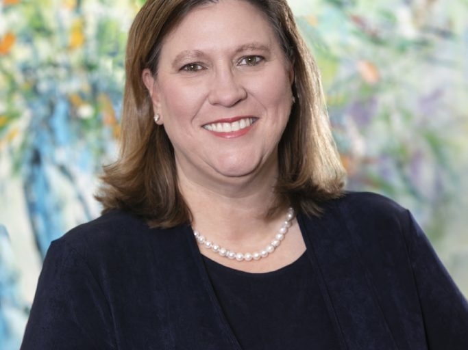 Julie Johnson is shown in this image wearing a dark outfit with a pearl necklace, smiling confidently in front of a vibrant, abstract background.