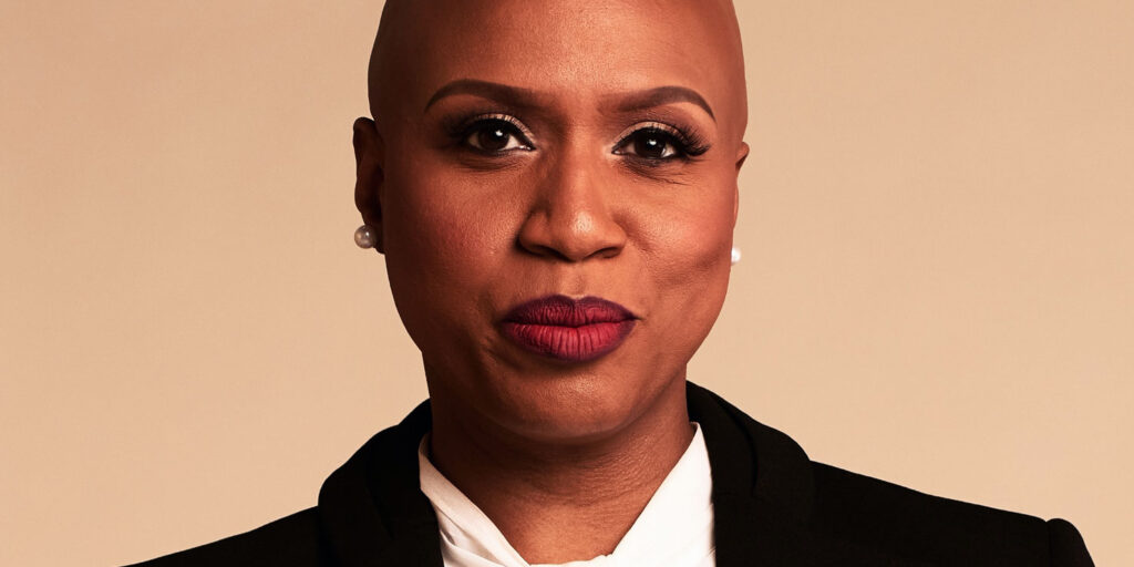 Ayanna Pressley is shown in this image wearing a black blazer and white blouse, with a confident expression, set against a neutral background.