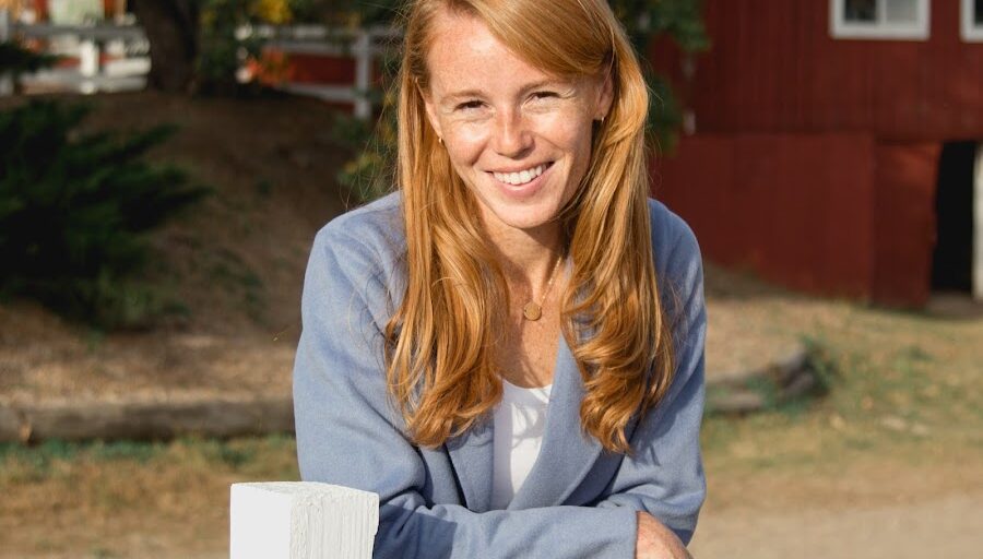 Rebecca Cooke smilling leaning over a fence with a barn-like background.