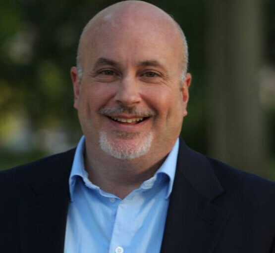 Mark Pocan is shown smiling outdoors in this image, wearing a light blue shirt and a dark blazer with a blurred green background.