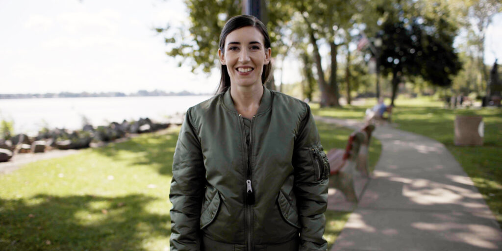 Ashley Ehasz is pictured outdoors in a green jacket, standing in a peaceful park setting with a serene water backdrop.