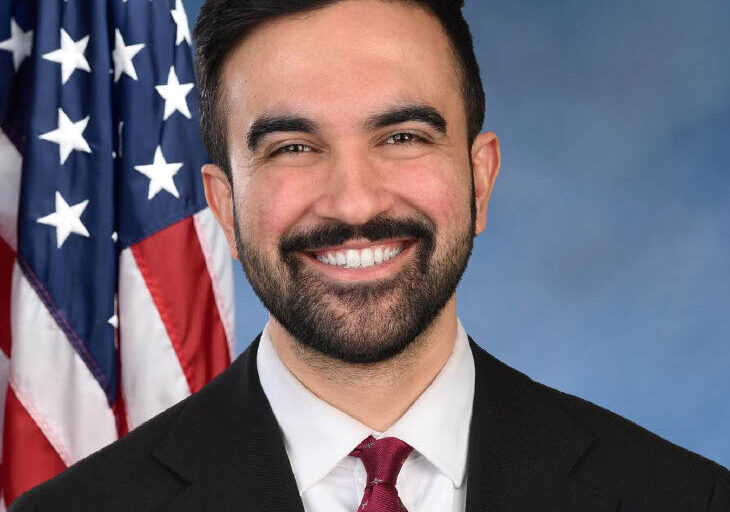 Zohran Mamdani for NYC Mayor portrait smiling at the camera wearing a black suit with red tie. He is posed with a blue background and an American flag behind his right shoulder.