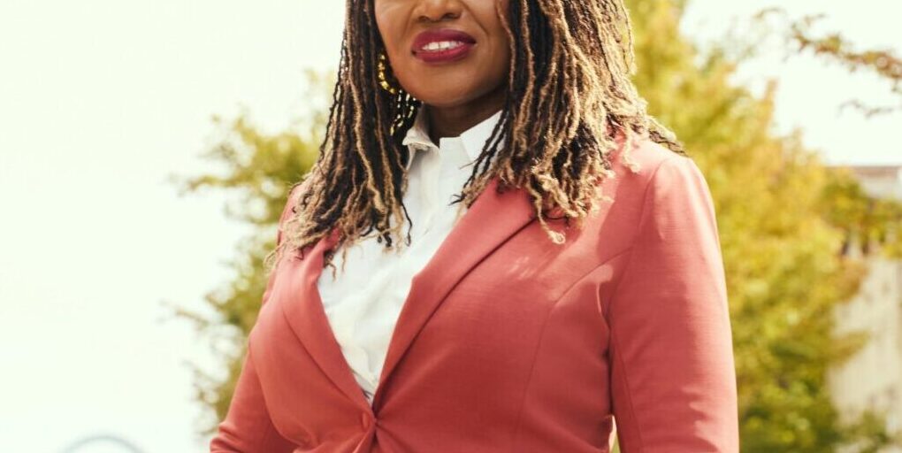 This is an image of Cori Bush, candidate for Missouri's 1st Congressional District. She is pictured in a red suit on a street in front of the St. Louis arch.