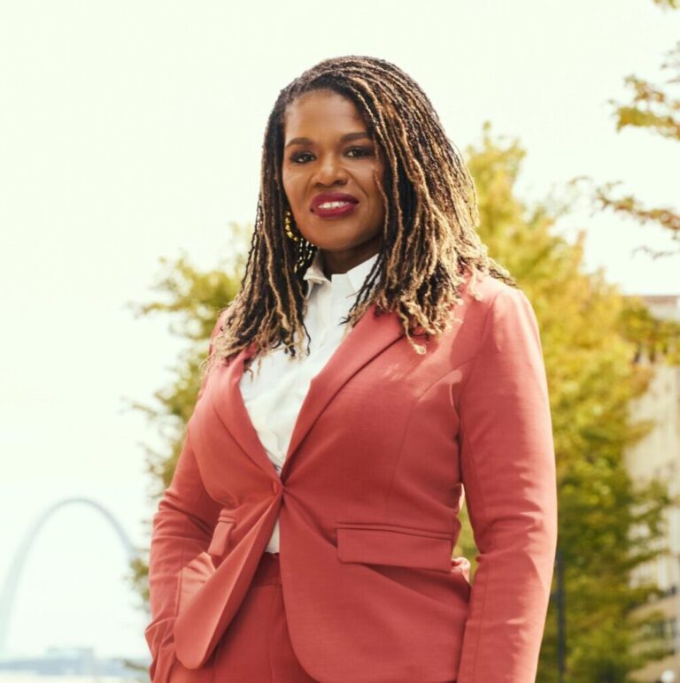 This is an image of Cori Bush, candidate for Missouri's 1st Congressional District. She is pictured in a red suit on a street in front of the St. Louis arch.