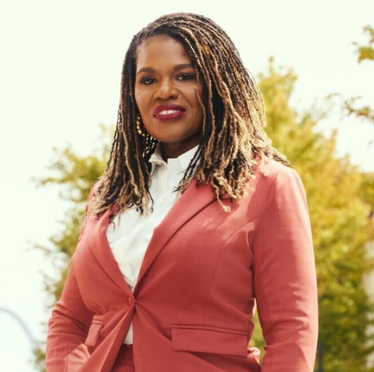 This is an image of Cori Bush, candidate for Missouri's 1st Congressional District. She is pictured in a red suit on a street in front of the St. Louis arch.