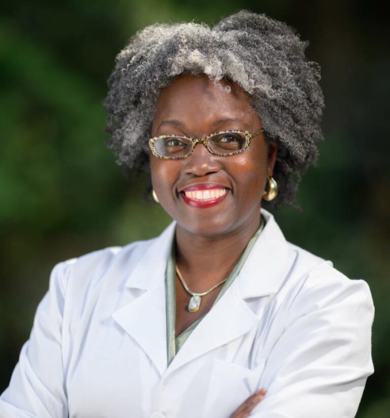 This is an image of Dr. Jasmine Clark, candidate for Georgia's 13th Congressional District. She is pictured in a white lab coat in front of trees.