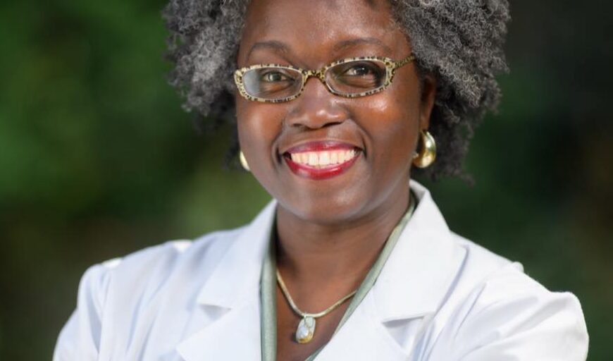 This is an image of Dr. Jasmine Clark, candidate for Georgia's 13th Congressional District. She is pictured in a white lab coat in front of trees.
