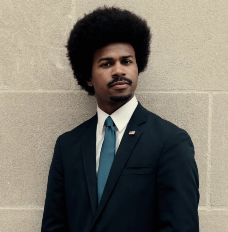 This is an image of Justin J. Pearson, candidate for Tennessee's 9th Congressional District. He is pictured in a black suit in front of a wall.