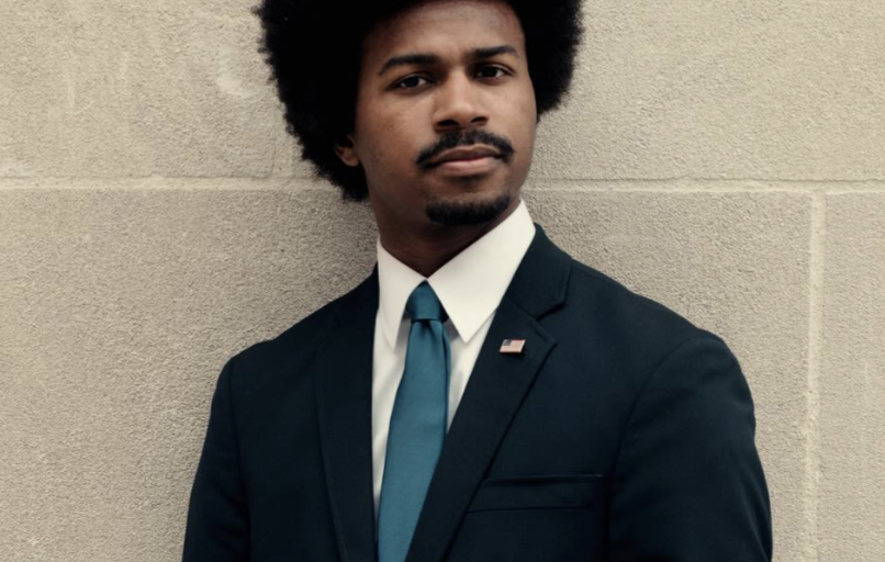 This is an image of Justin J. Pearson, candidate for Tennessee's 9th Congressional District. He is pictured in a black suit in front of a wall.
