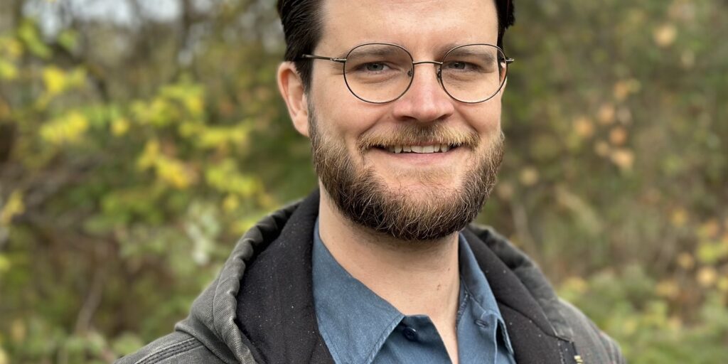 This is an image of William Lawrence, candidate for Michigan's 7th Congressional District. He is pictured in a blue shirt and gray hoodie in front of a forest.