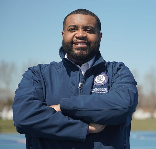 This is an image of Donavan McKinney, candidate for Michigan's 13th Congressional District. He is pictured in a blue jacket in front of a basketball court in a park.