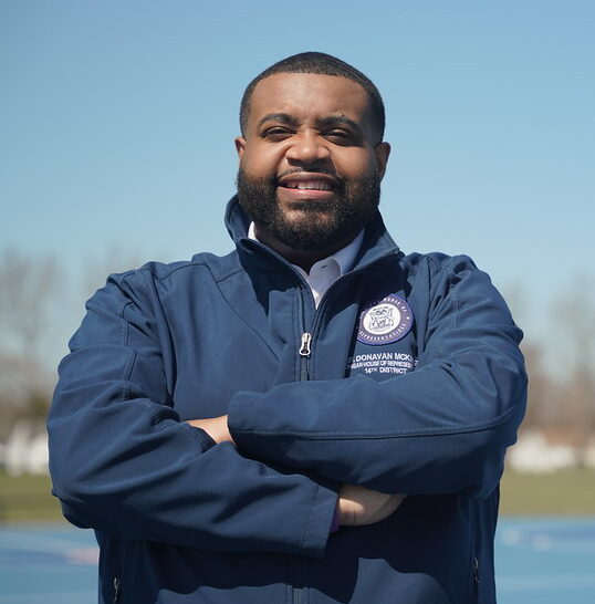 This is an image of Donavan McKinney, candidate for Michigan's 13th Congressional District. He is pictured in a blue jacket in front of a basketball court in a park.