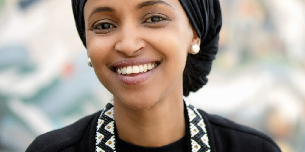 This is an image of Ilhan Omar, candidate for Minnesota's 5th Congressional District. She is pictured in a black shirt with a black hijab standing in front of a street mural.