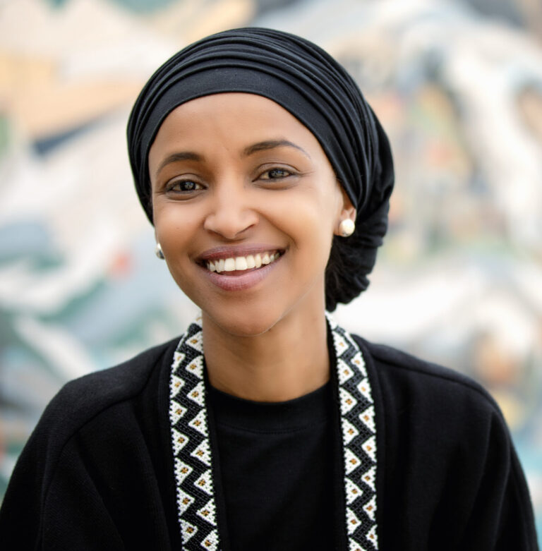 This is an image of Ilhan Omar, candidate for Minnesota's 5th Congressional District. She is pictured in a black shirt with a black hijab standing in front of a street mural.