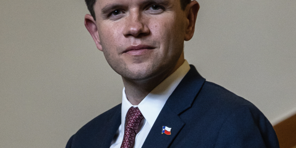 This is an image of James Talarico, candidate for Texas Senate. He is pictured in a blue suit with a white shirt and red tie standing on a brown staircase.