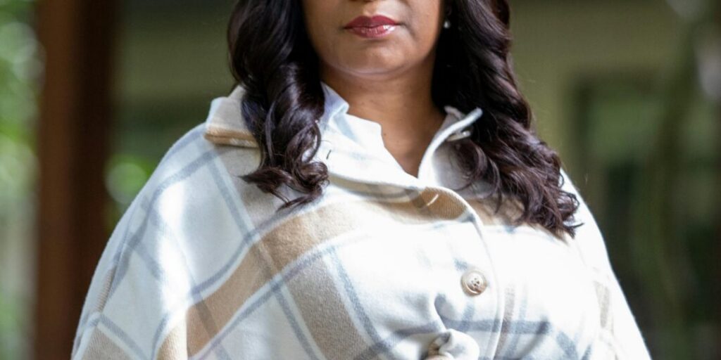 This is an image of Janelle Bynum, candidate for Oregon's 5th Congressional District. She is pictured in a white coat with brown stripes standing in front of house.
