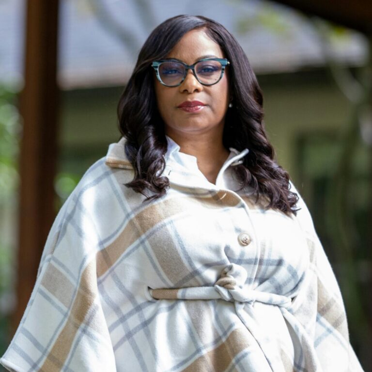 This is an image of Janelle Bynum, candidate for Oregon's 5th Congressional District. She is pictured in a white coat with brown stripes standing in front of house.