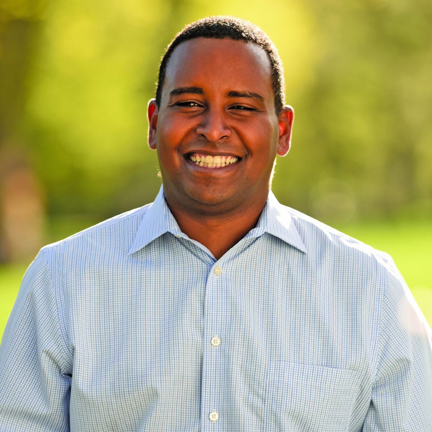 This is an image of Joe Neguse, candidate for Colorado's 2nd Congressional District. He is pictured in a blue shirt standing with a blurred background of trees.