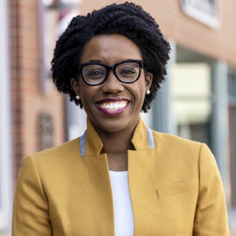 This is an image of Lauren Underwood, candidate for Illinois's 14th Congressional District. She is pictured in a dark yellow jacket in front of a buidling.