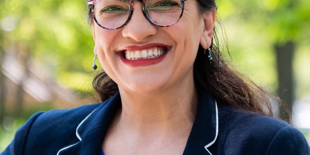 This is an image of Rashida Tlaib, candidate for Michigan's 12th Congressional District. She is pictured in a navy blue jacket with a blue shirt standing in front of trees.