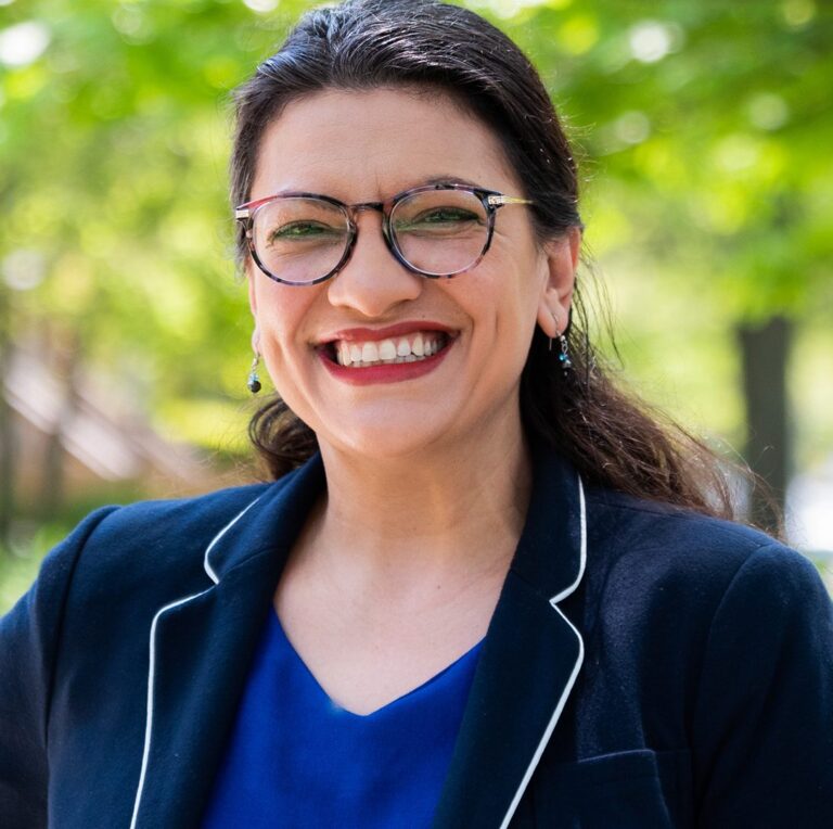 This is an image of Rashida Tlaib, candidate for Michigan's 12th Congressional District. She is pictured in a navy blue jacket with a blue shirt standing in front of trees.
