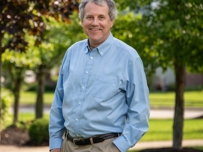 This is an image of Sherrod Brown, candidate for Ohio Senate. He is pictured in a blue shirt standing in front of trees in front of a road.