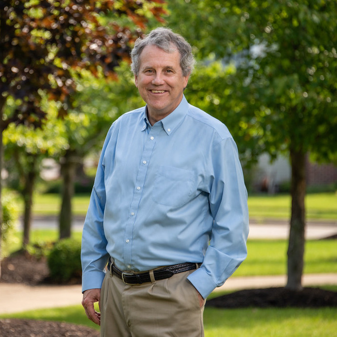This is an image of Sherrod Brown, candidate for Ohio Senate. He is pictured in a blue shirt standing in front of trees in front of a road.
