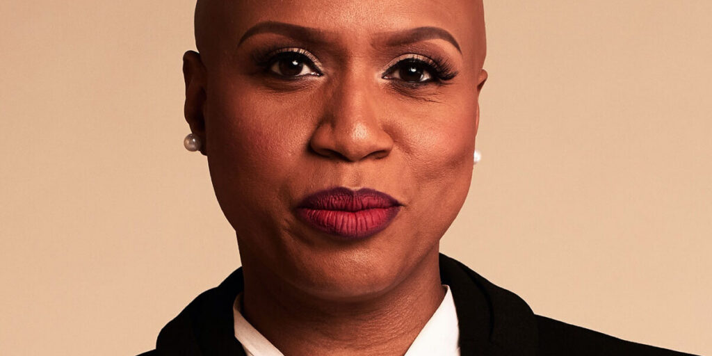This is an image of Ayanna Pressley, candidate for Massachusetts's 7th Congressional District. She is pictured in a black blazer with a white shirt in front of a bronze background.