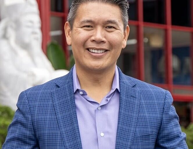 This is an image of Derek Tran, candidate for California's 45th Congressional District. He is pictured in a blue suit with a purple shirt standing in front a red building.