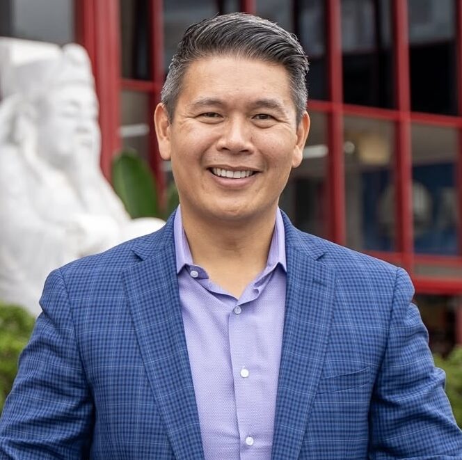 This is an image of Derek Tran, candidate for California's 45th Congressional District. He is pictured in a blue suit with a purple shirt standing in front a red building.