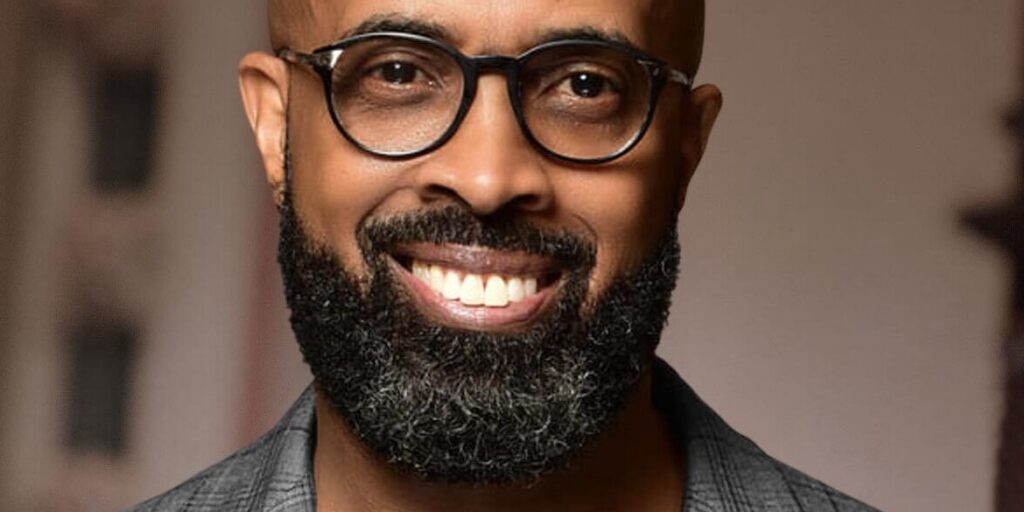 This is an image of Rev. Frederick D. Haynes III, candidate for Texas's 30th Congressional District. He is pictured in a striped gray suit with a black shirt in front of a brown background.