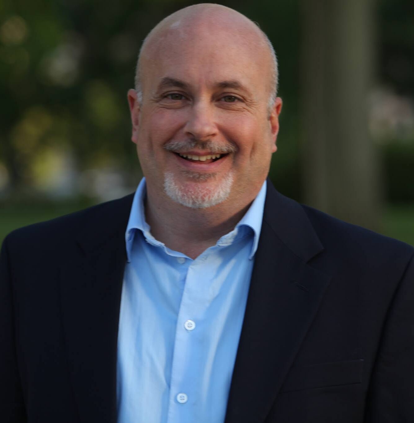This is an image of Mark Pocan, candidate for Wisconsin's 2nd Congressional District. He is pictured in a black suit with a light blue shirt standing in front a tree.