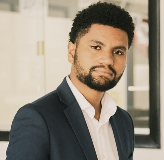 This is an image of Maxwell Alejandro Frost, candidate for Florida's 10th Congressional District. He is pictured in a black suit with a white shirt standing in front of a large mirror.