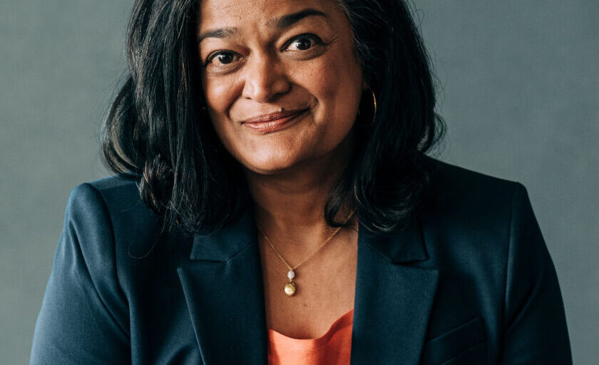 This is an image of Pramila Jayapal, candidate for Washington's 7th Congressional District. She is pictured in a black suit with a peach shirt in front of a gray wall.