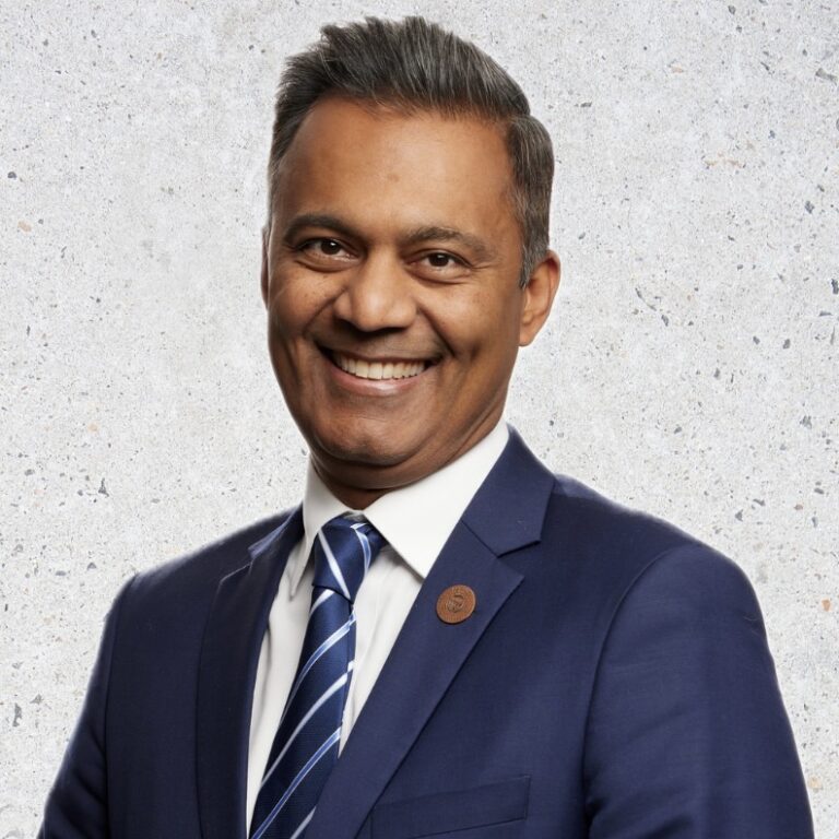 This is an image of Amish Shah, candidate for Arizona's 1st Congressional District. He is pictured in a blue suit standing in front of a white background.