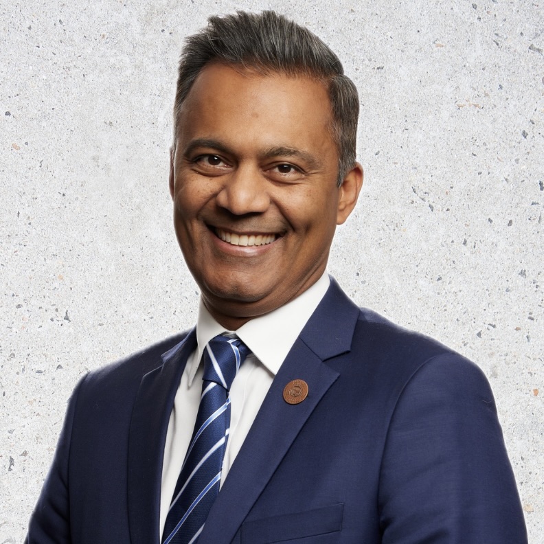 This is an image of Amish Shah, candidate for Arizona's 1st Congressional District. He is pictured in a blue suit standing in front of a white background.